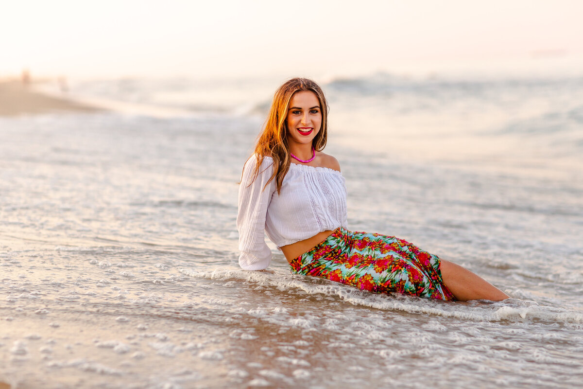 A high school senior lounges effortlessly in the surf at golden hour, wearing a colorful floral skirt and off-the-shoulder white blouse.  Captured by Brooke Tucker Photography in Virginia Beach, this portrait celebrates confidence, personality, and the wild beauty of the sea. 