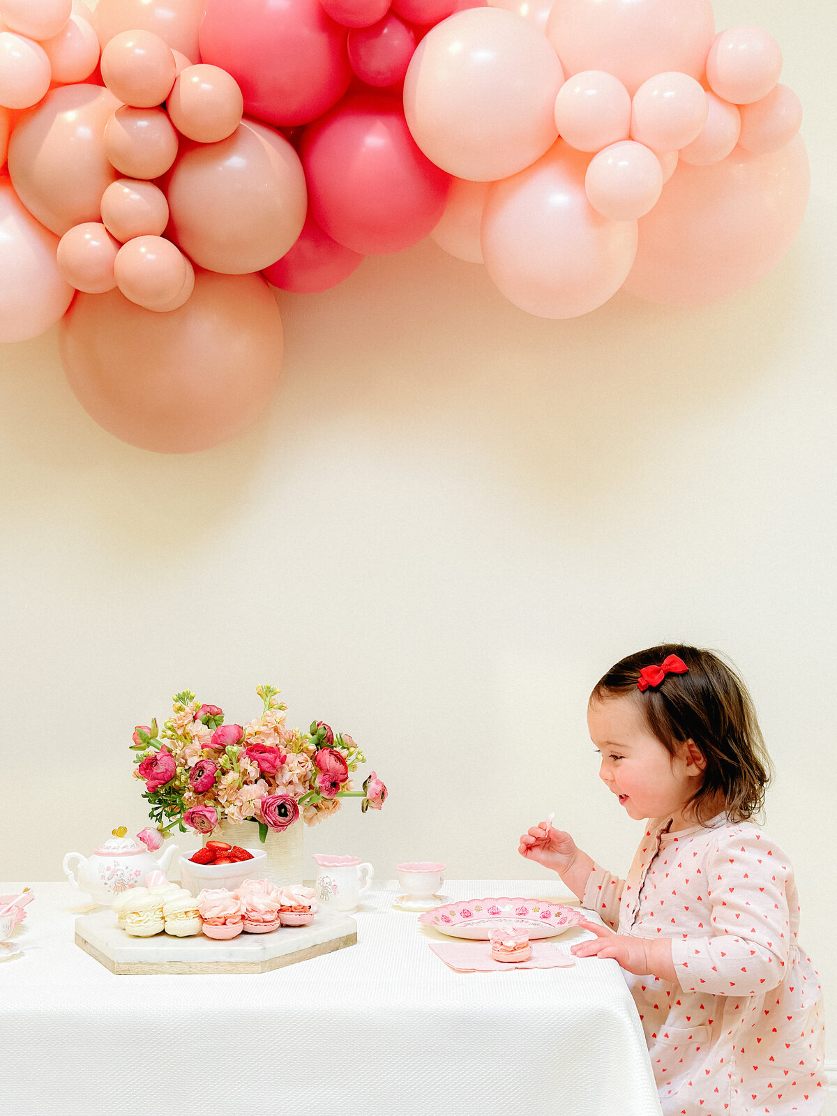 Little girl at a table with flowers and cake underneath a balloon arch