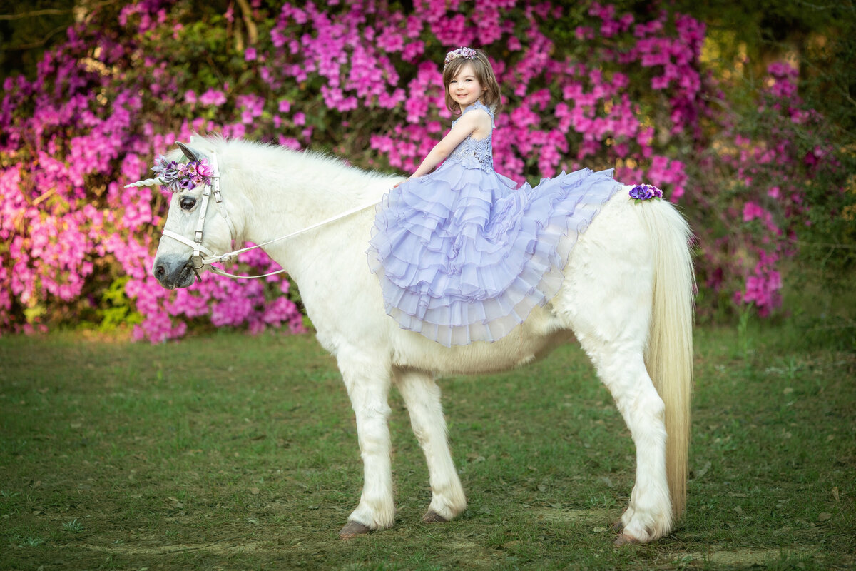 Young girl on white horse with unicorn horn.  The girl is wearing a periwinkle dress from Butterfly Closet and there are pink azaleas in the background.  The girl is sitting on the Welsh pony, holding the reins, and looking sideways towards the camera.