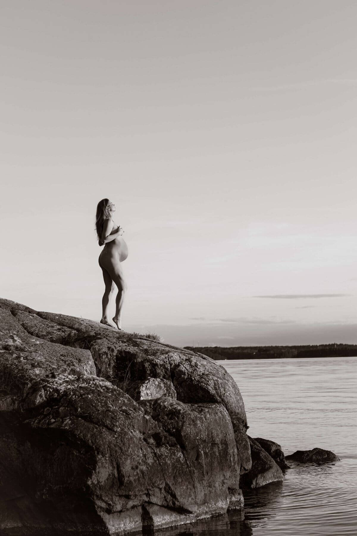 Gravidbild med naken kvinna ståendes på ett berg ute vid vattnet under en gravidfotografering i Stockholm