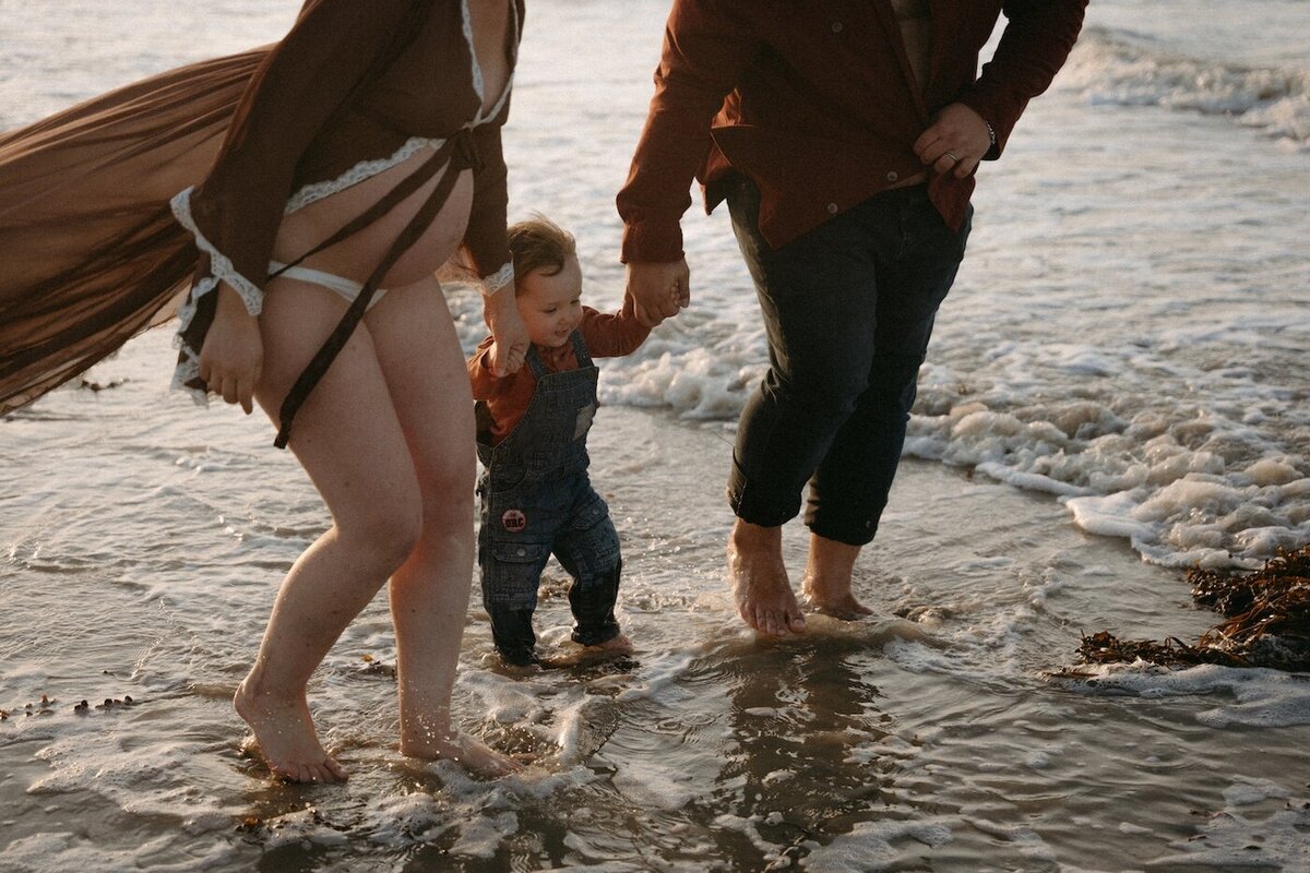 Couple marchant pieds nus dans l'eau avec leur petit garçon qu'ils tiennent par les mains. Par Kamescop, reportage vidéo et photo famille en Bretagne.