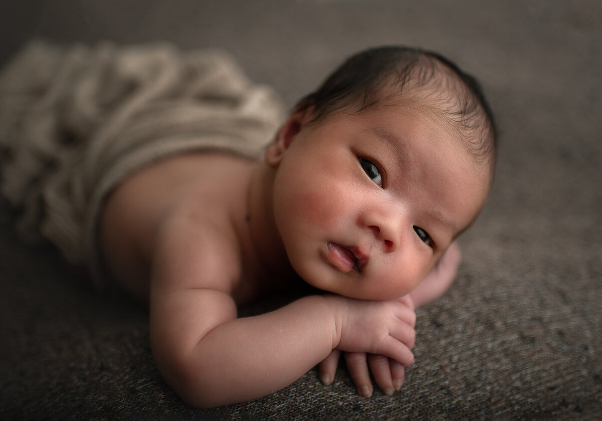 newborn looking at camera while laying on hands and brown blanket at newborn session