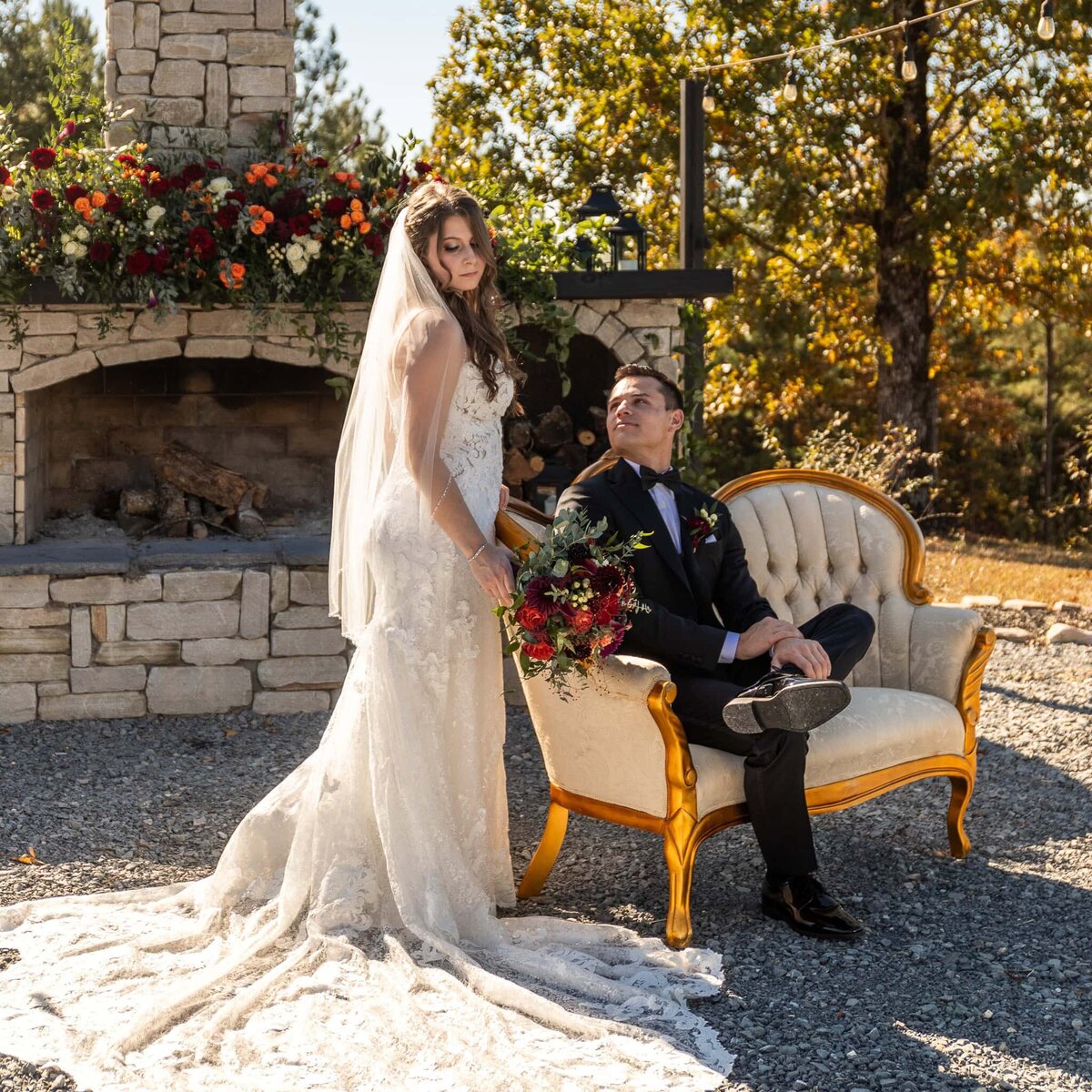 The groom sits on a champagne colored couch outdoors in front of a decorated stone fireplace and looks at his bride who is standing beside him, holding her bouquet of red and purple flowers, looking down her right shoulder.