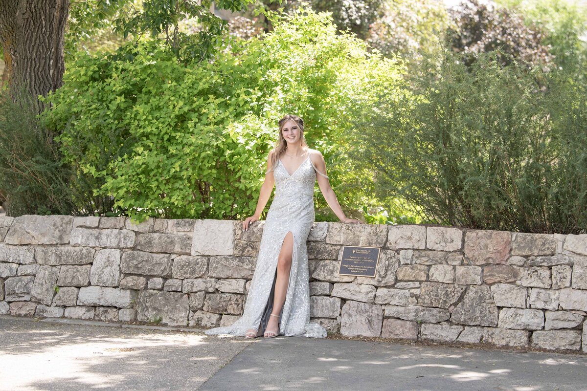 Graduate in a silver dress leaning against a stone wall in Saskatoon.