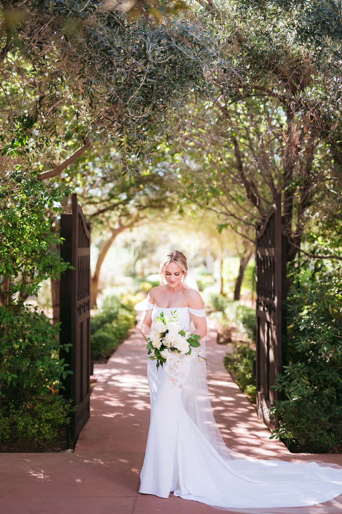 Bride standing beneath the garden archway at El Chorro in Scottsdale, holding a white floral bouquet in elegant natural light.