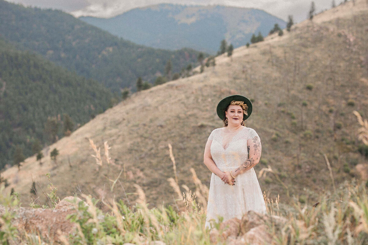 The bride, with an emerald brimmed hat and lacy boho gown, smiles for the camera in front of a mountain overlook  in Colorado