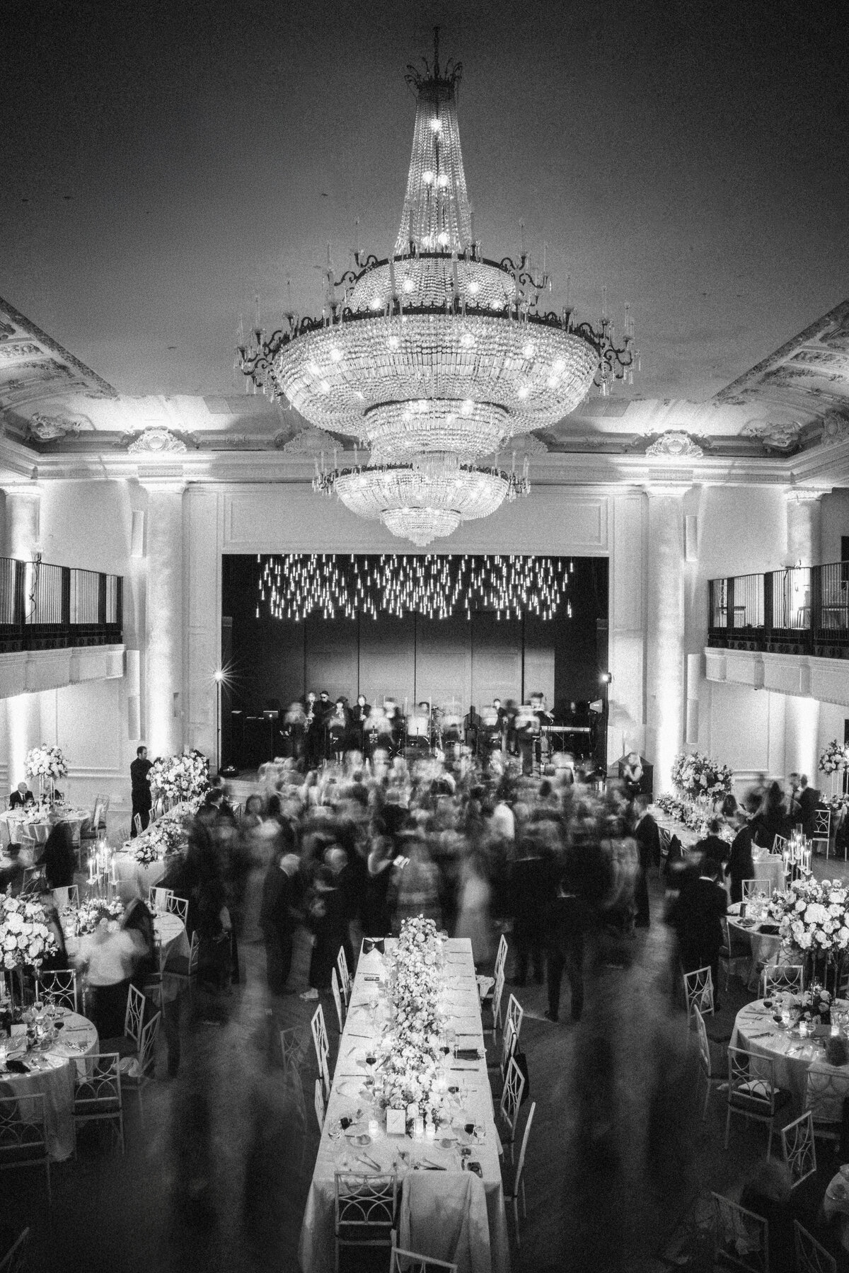 Black and white photo of a wedding reception in a grand ballroom with a towering crystal chandelier, blurred motion of guests dancing, long banquet tables adorned with floral arrangements, and a live band performing on a raised stage in the background.