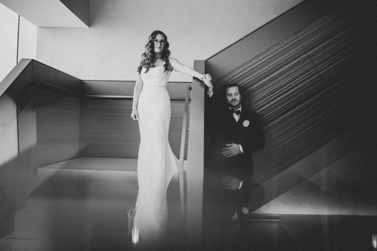A black-and-white portrait of a couple holding hands during their first touch standing apart from each other on a modern staircase, capturing an elegant and timeless wedding moment.