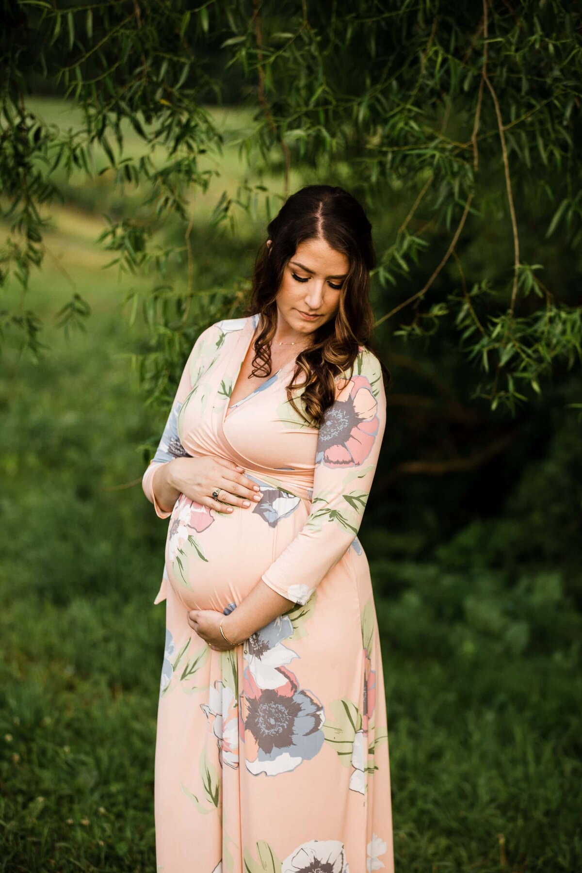Pregnant woman in a floral dress holding her belly under a tree for her maternity photography session.