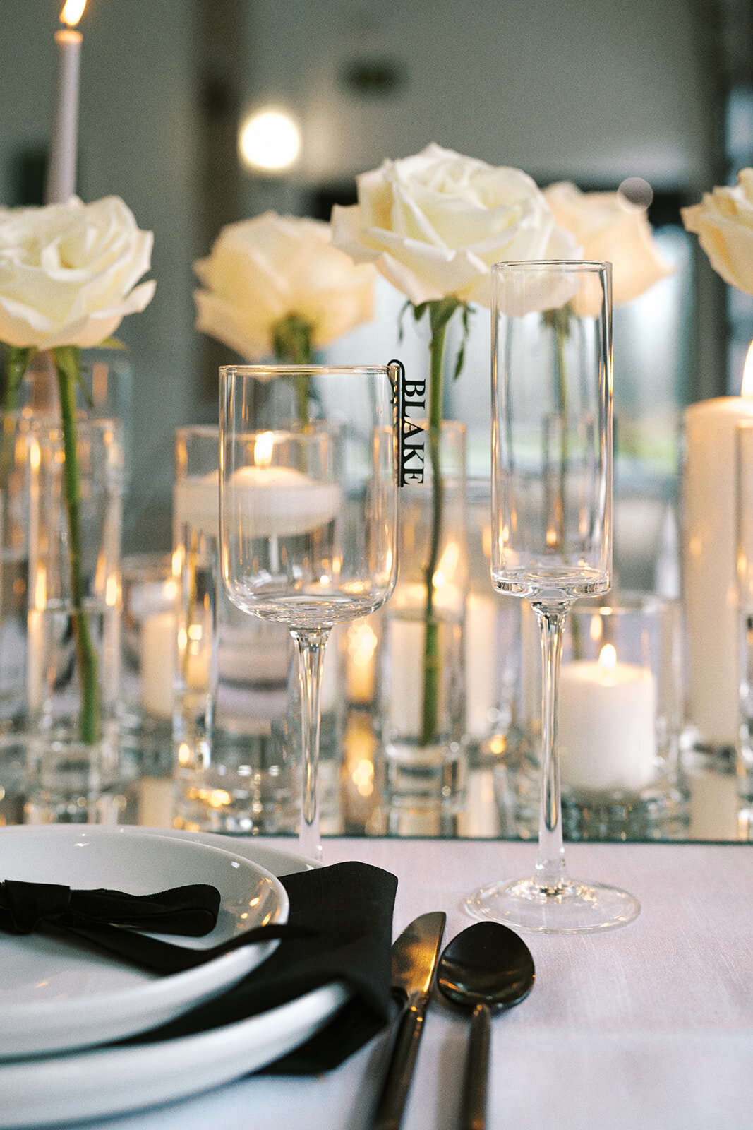 A close up of glassware, lit candles and white roses on a table at a styled wedding photography shoot.