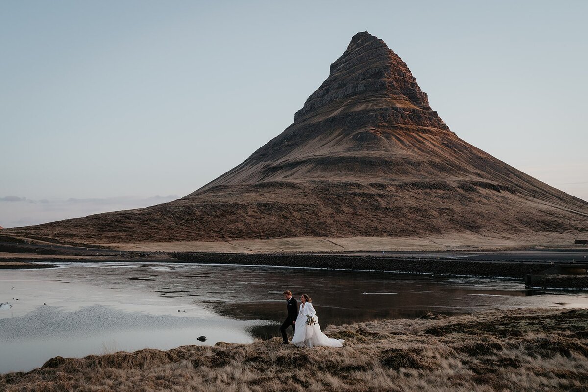 Bride and groom hold hands while walking in front of a lake and mountain during their elopement in Iceland. 