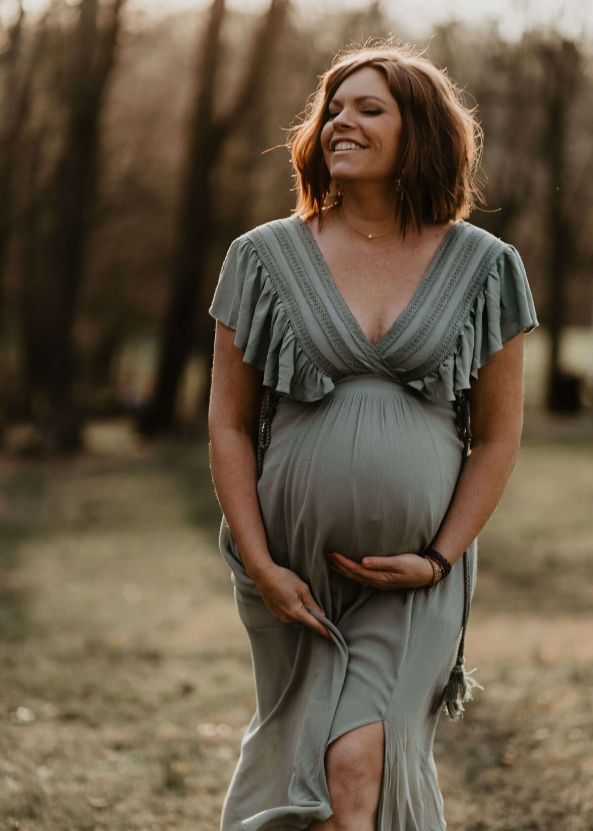 A pregnant woman in a sage green dress is strolling through the woods, captured by a Pittsburgh maternity photographer.