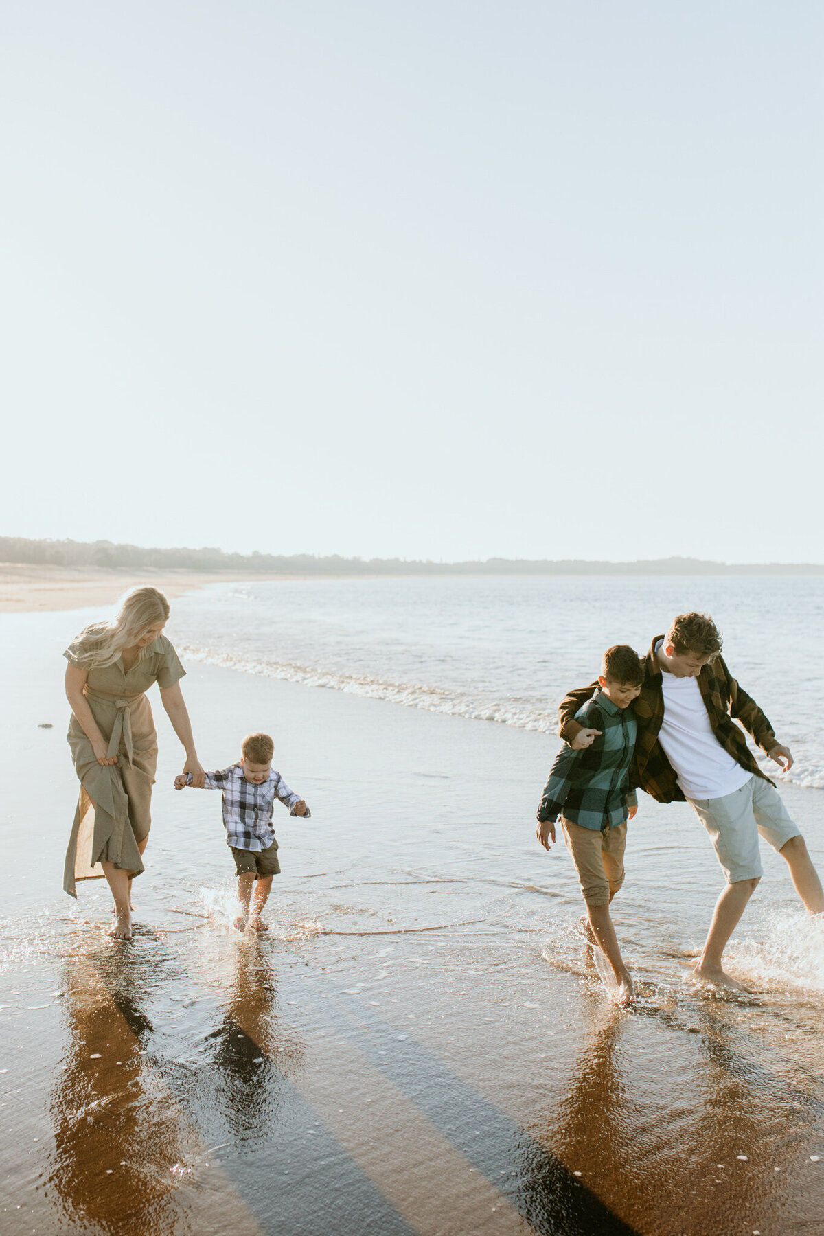 Family of four walking along the beach, kicking water as the sun casts a soft glow behind them.