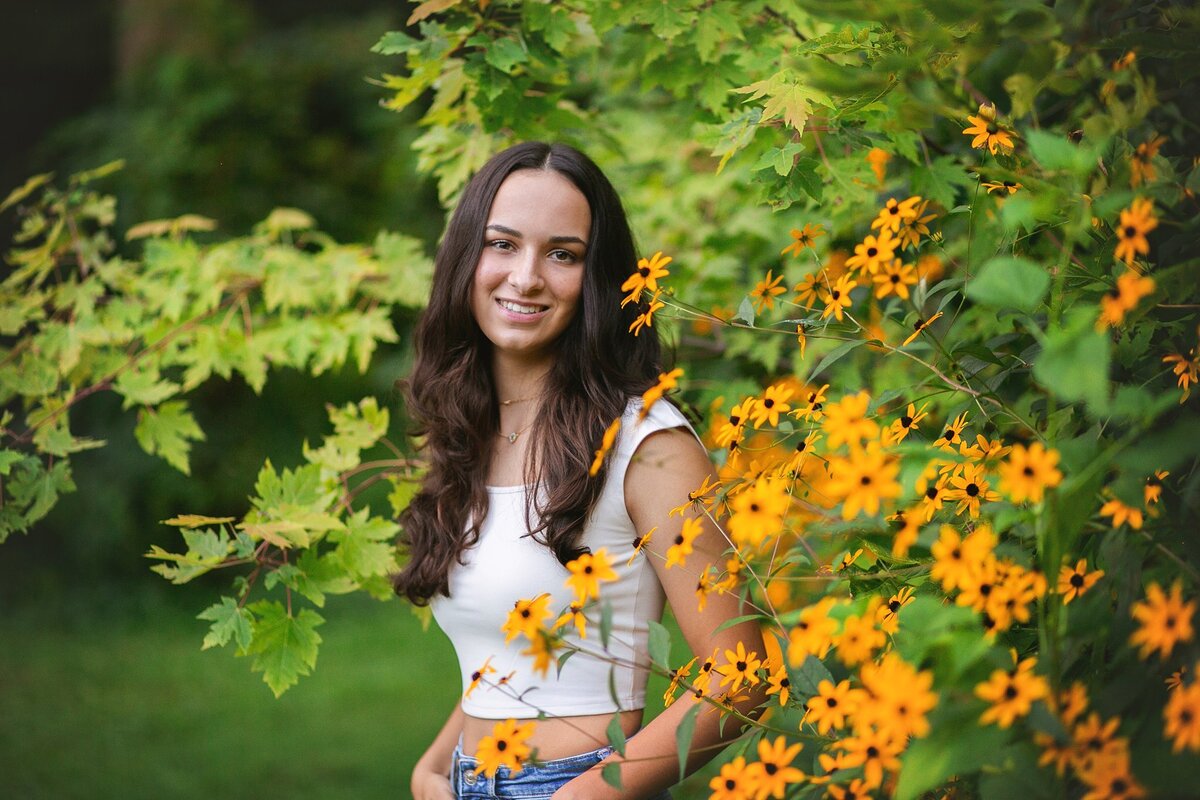 high-school-senior-with-cropped-white-tee-and-blue-jeans-leans-in-field-of-flowers-at-100-acre-woods-on-west-side-of-downtown
