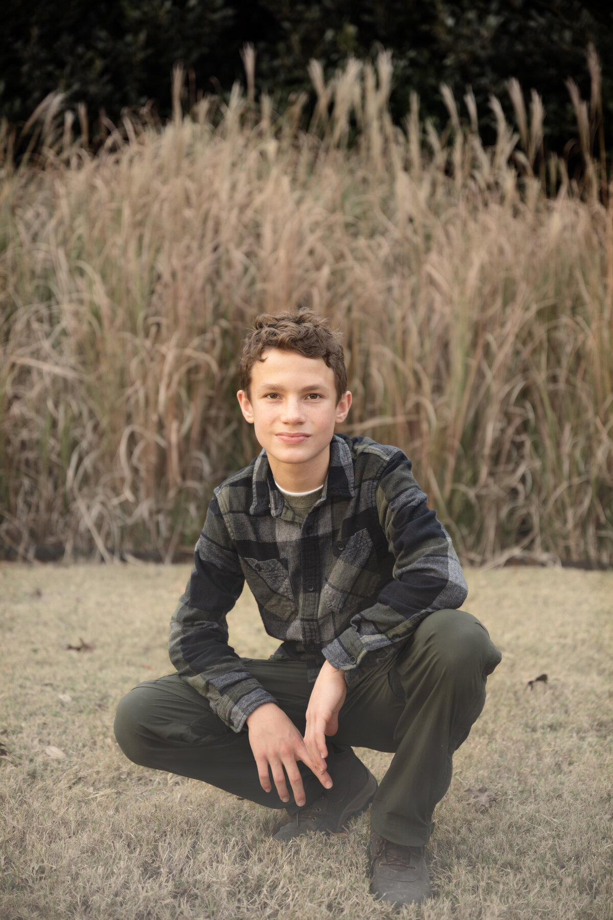 A young boy wearing flannel poses in front of tall grass on a foggy morning during the family's outdoor family photo session in McKinney, TX.