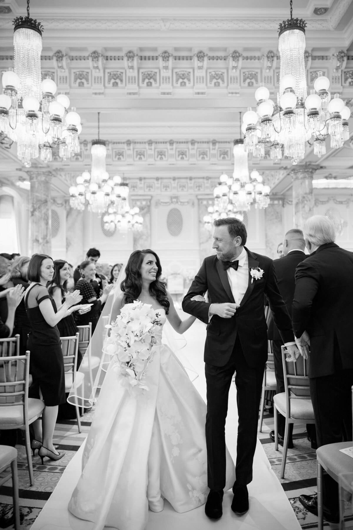 A bride and groom walk hand in hand down the aisle in an elegant venue. The bride wears a long, flowing gown and holds a bouquet, while guests seated on either side applaud. The room is decorated with grand chandeliers and ornate details.
