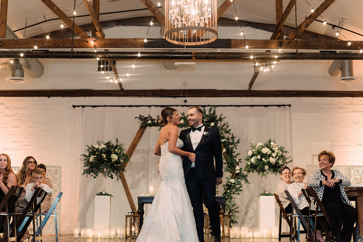 The bride and groom share a dance at their wedding reception, smiling and holding each other amidst an elegantly decorated venue with string lights and floral arrangements, while guests watch with smiles.
