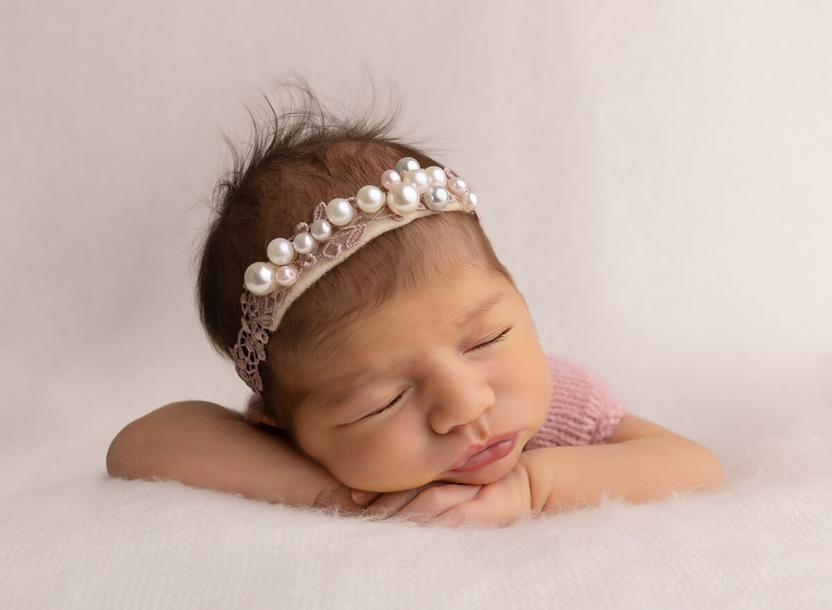 Baby girl in a pearl headband is sleeping for her brooklyn newborn photoshoot with her hands folded underneath her chin. Captured by top Brooklyn newborn photographer Chaya Bornstein