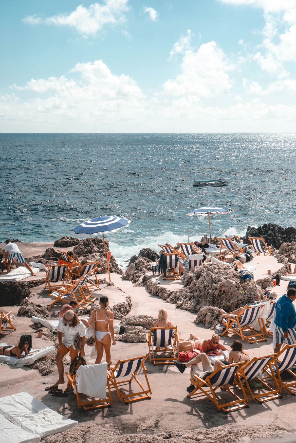 People relaxing on a rocky beach with blue and white striped deck chairs and umbrellas. The ocean is visible in the background under a partly cloudy sky. Embracing the European travel specialist ambiance, they are sunbathing, talking, and enjoying the seaside atmosphere.