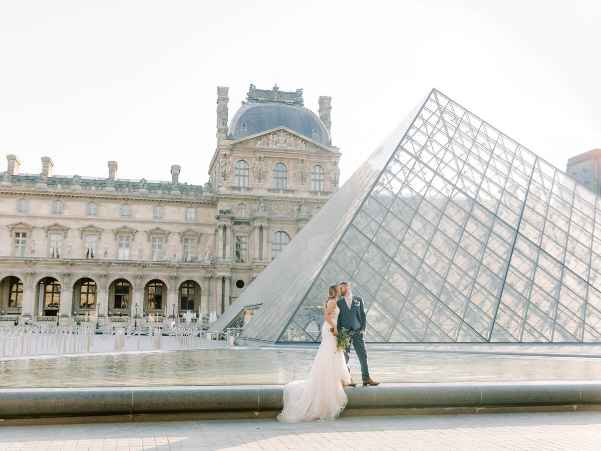 A bride and groom standing on the edge of Louvre  fountain with glass pyramid at the background