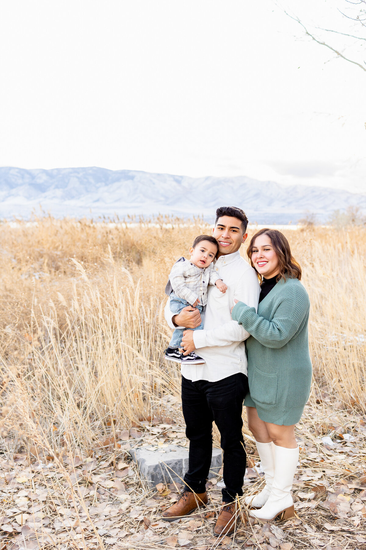 A young couple stands in a field of tall golden grass with their toddler, smiling warmly at the camera with mountains stretching across the horizon behind them.
