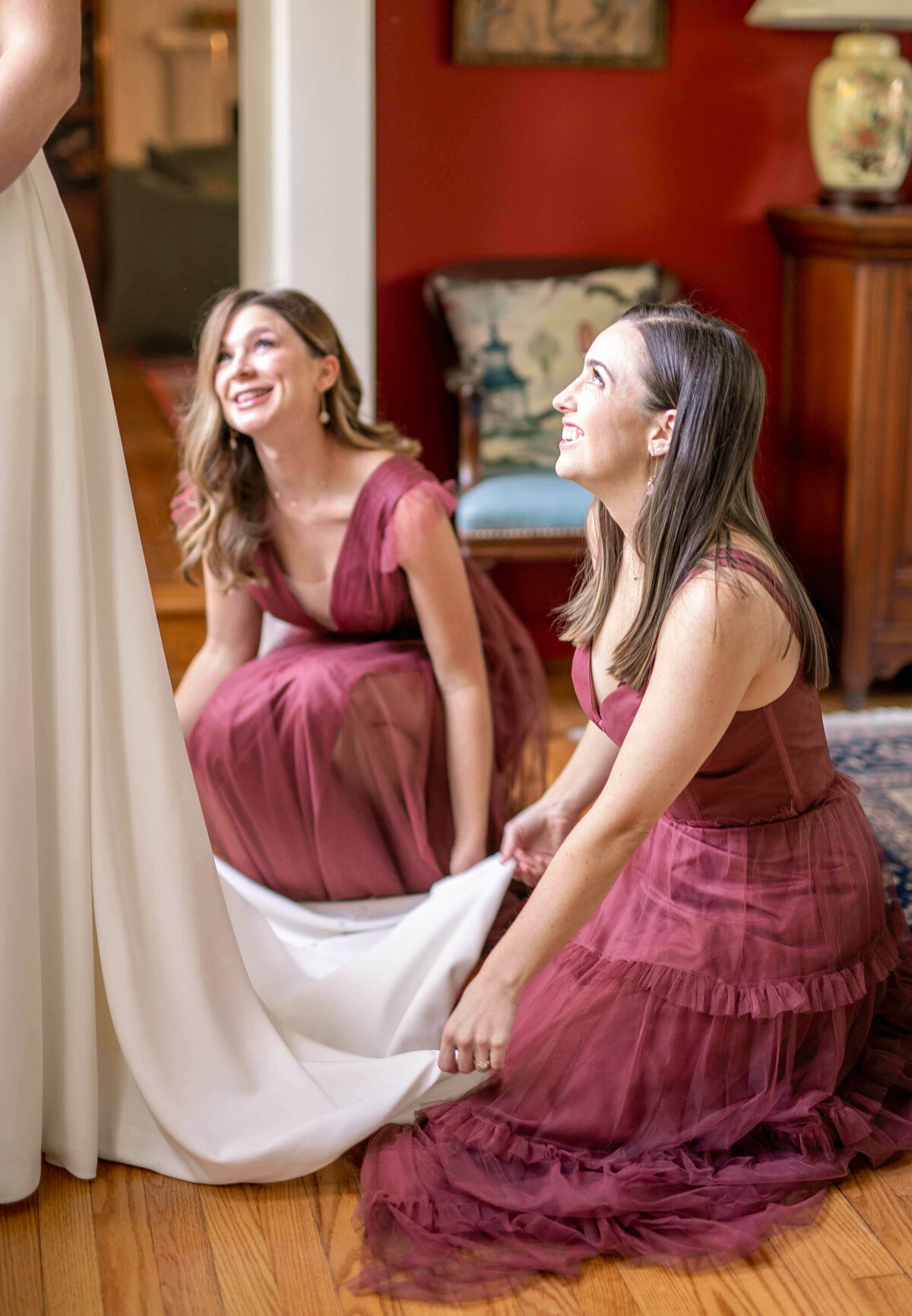 Two women in burgundy dresses smiling and kneeling, adjusting the train of a white dress. They appear joyful, surrounded by warm home decor, including wooden furniture and a blue upholstered chair.