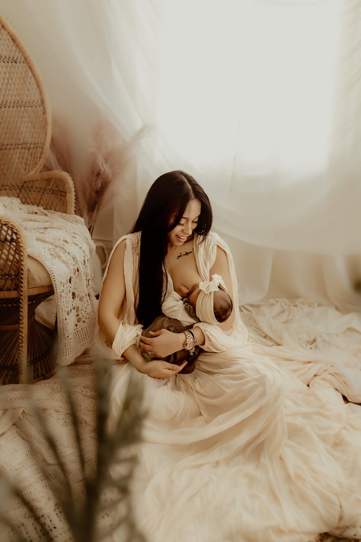 A woman in a light dress sits on a soft, fabric-covered floor, holding a swaddled baby. The softly lit setting highlights the expert touch of a Macon studio newborn photographer, with a wicker chair and draped curtains adding to the serene ambiance.