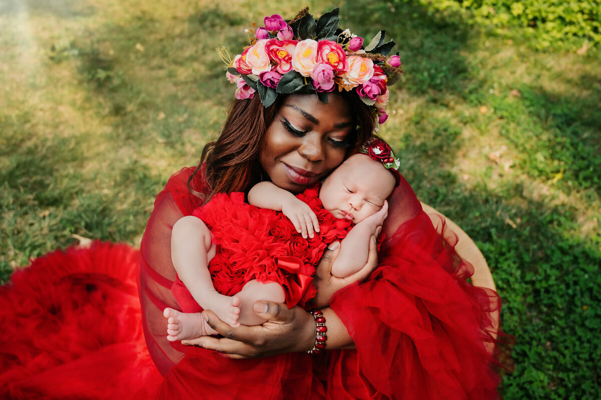 Mom wearing red gown and floral crown holding and looking down at her baby girl outdoors.