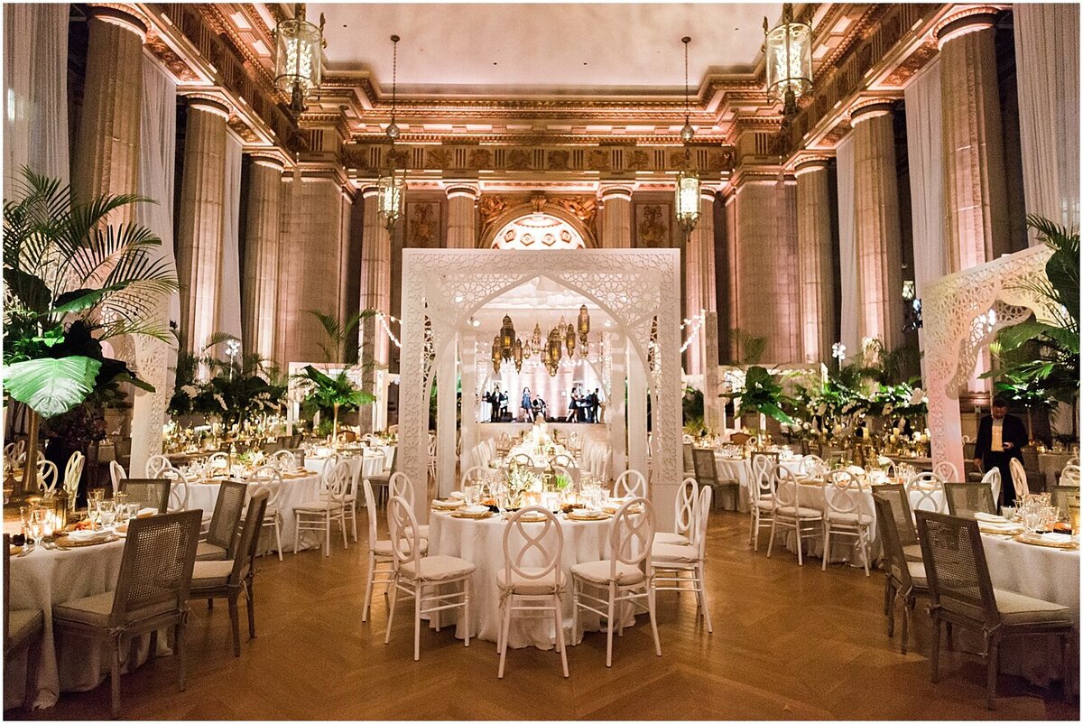 This is where the photo was taken at Washington D.C., The Mellon Auditorium, one of the best luxury DC venues with European flair, captured by photographer Sarah Bradshaw. The grand ballroom is elaborately decorated for an event with white tables and chairs arranged around an intricately designed white canopy. The room features tall columns, lush green plants, ornate lighting, and a beautifully illuminated ceiling.