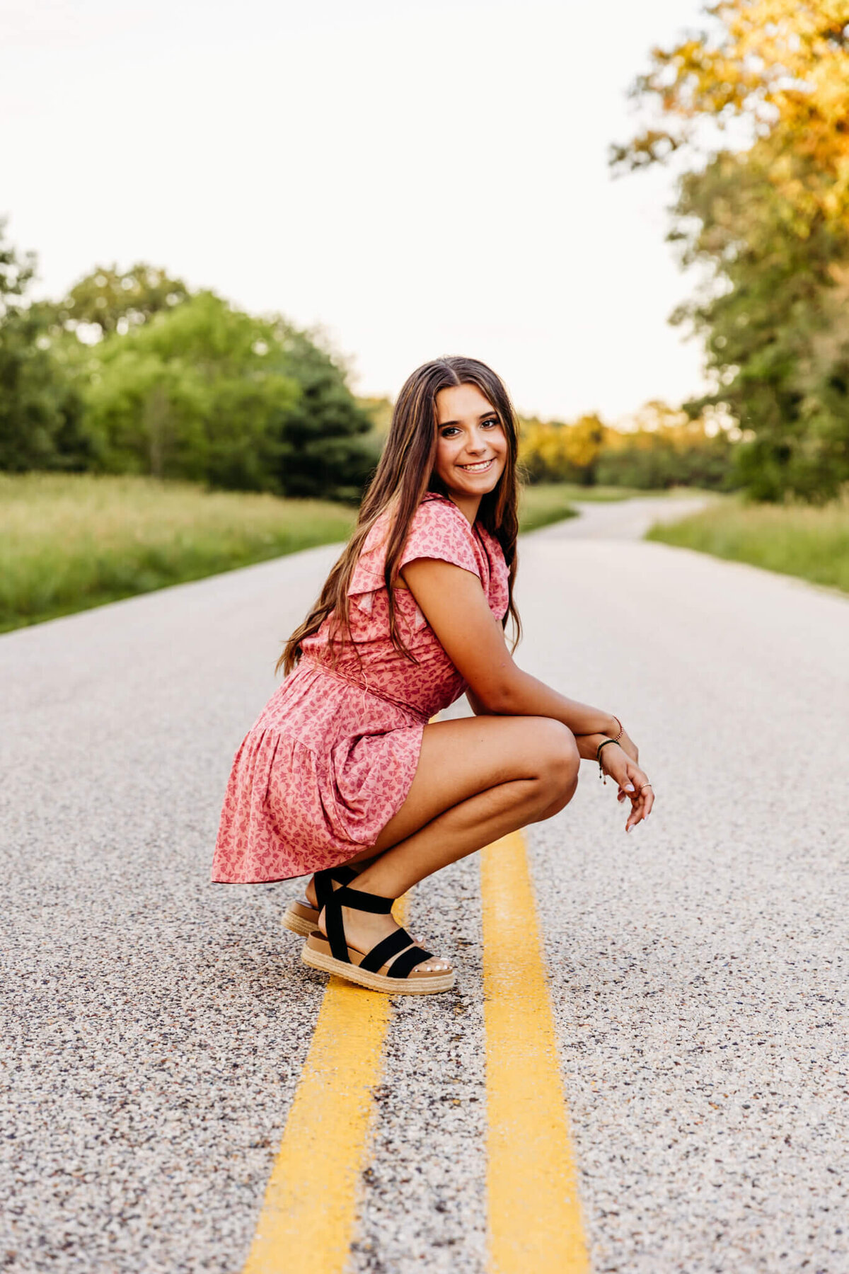 pretty teenage girl from an Oshkosh high school sitting on the yellow lines of a back road and resting her hands on her knees as she smiles and looks over her shoulders captured by Ashley Kalbus