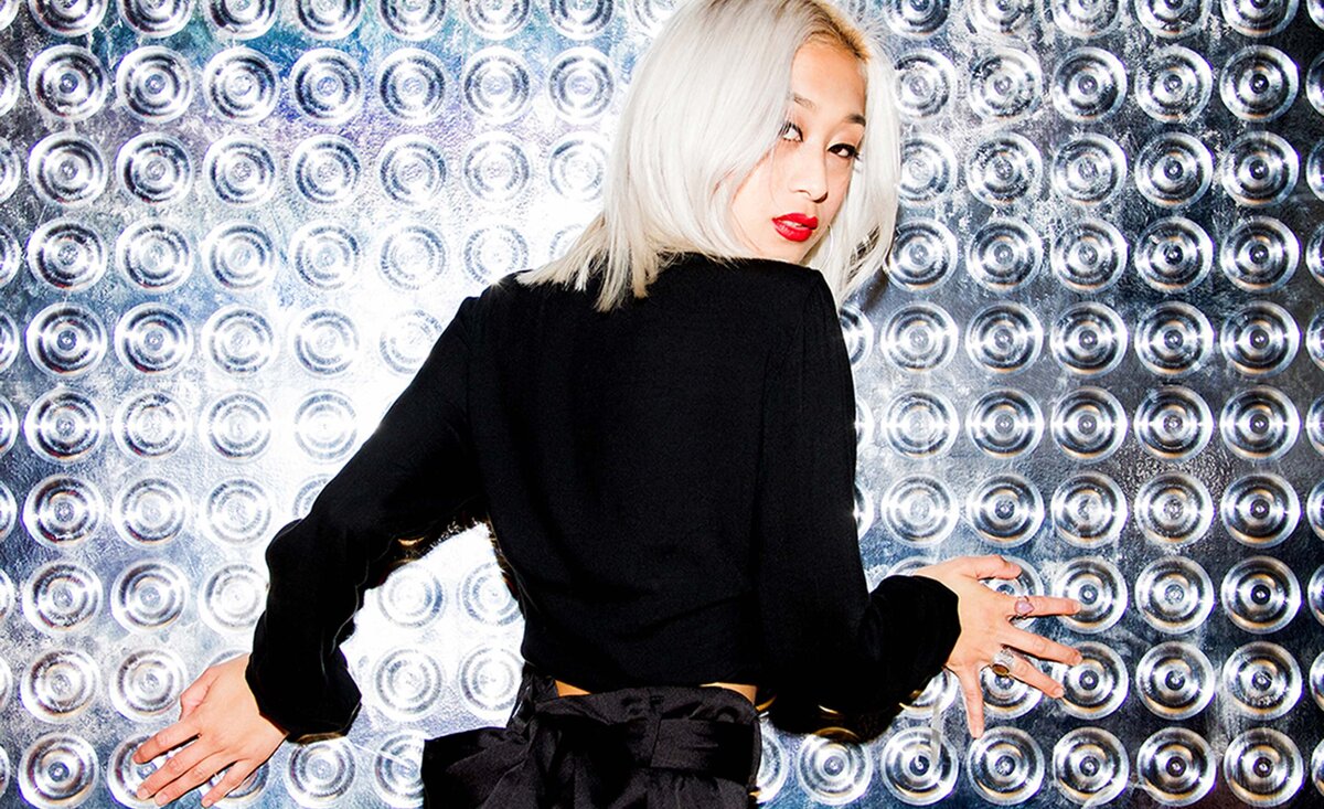 Female musician portrait Wynona Hulipas wearing black dress leaning against silver circle  wall looking back over her shoulder