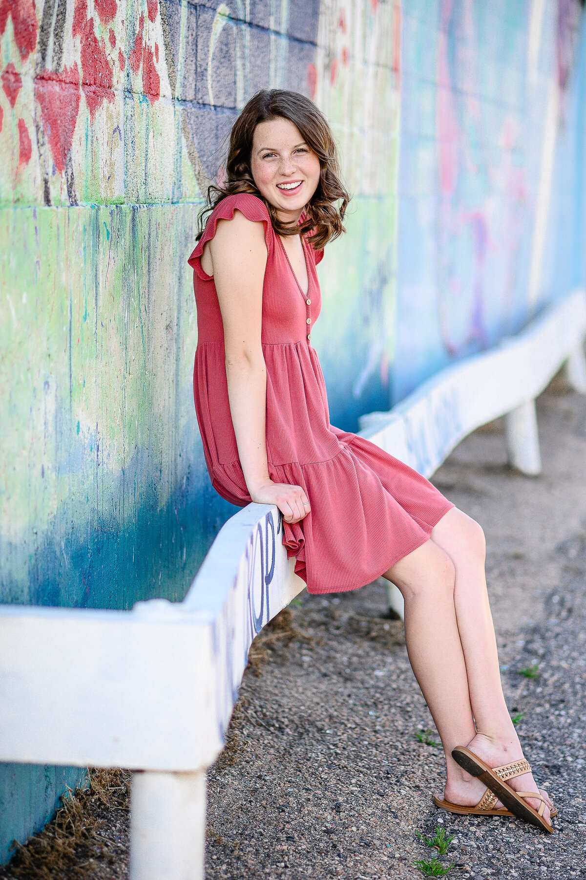 senior picture photographers near me  captures senior picture dresses with girl wearing a pink sun dress sitting on a wooden fence next to wall art in Denver