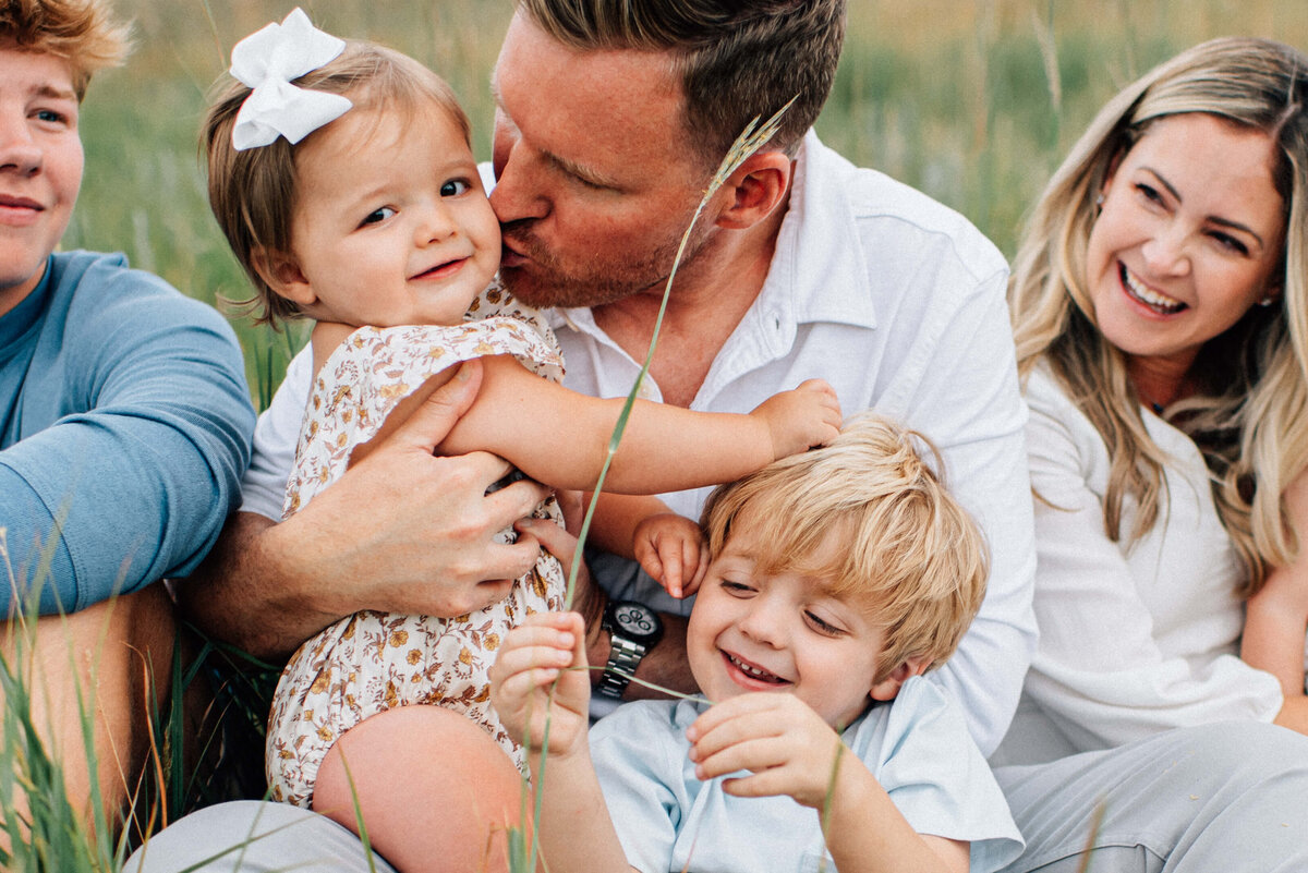 family sitting and snuggling in grass with dad kissing baby daughter