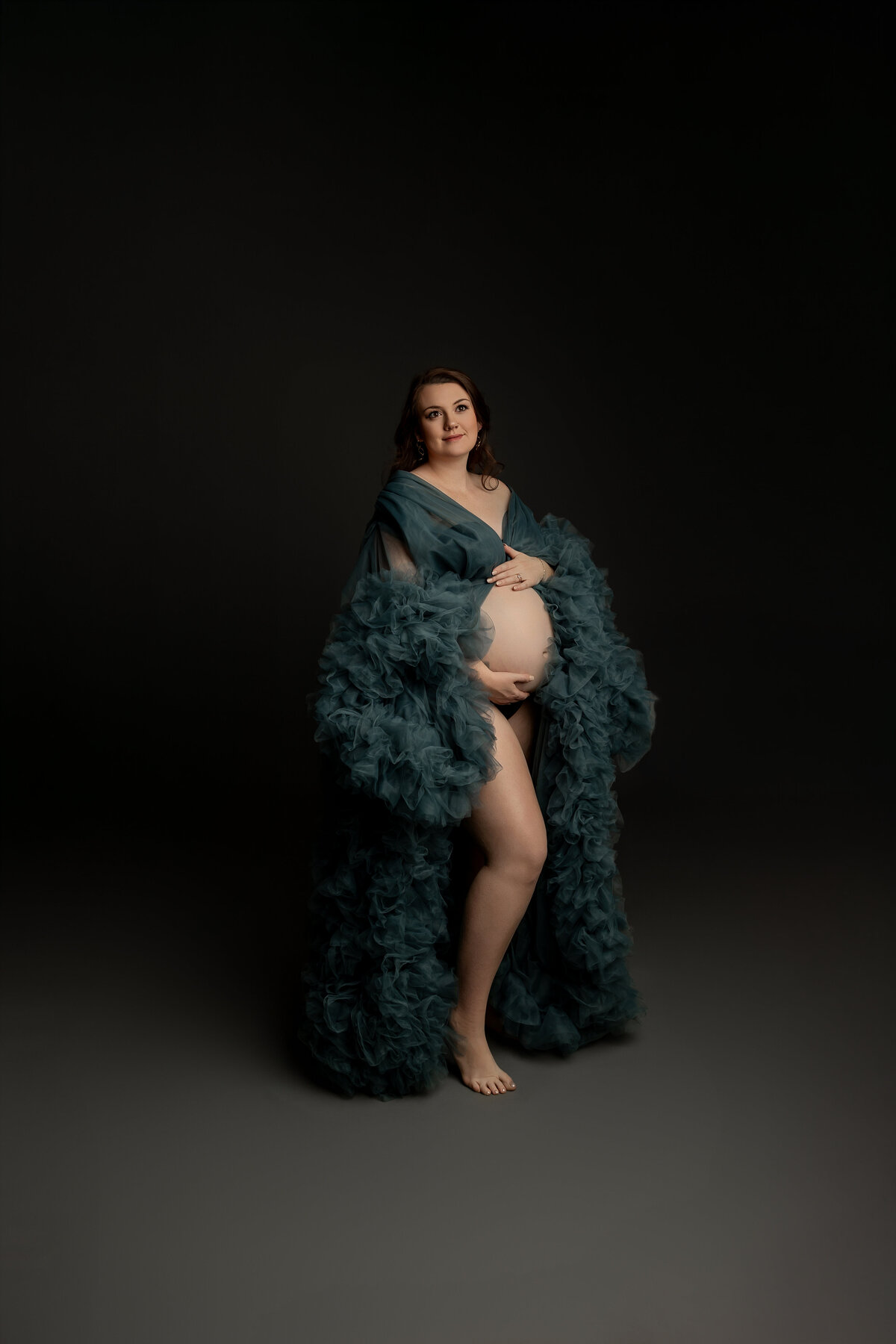 Wearing a graceful green dress, the expectant mother poses in a side view, her hands gently resting on her belly as she radiates calm and anticipation. Captured by Inland Empire maternity photographer Kaitlyn Dawn Photography.