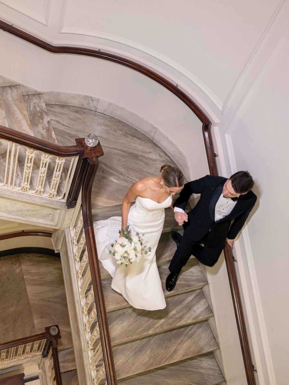 A bride in a white gown and a groom in a black tuxedo walk hand in hand down a curved staircase. The bride holds a bouquet as they descend the ornate steps with wooden railings and marble floors.