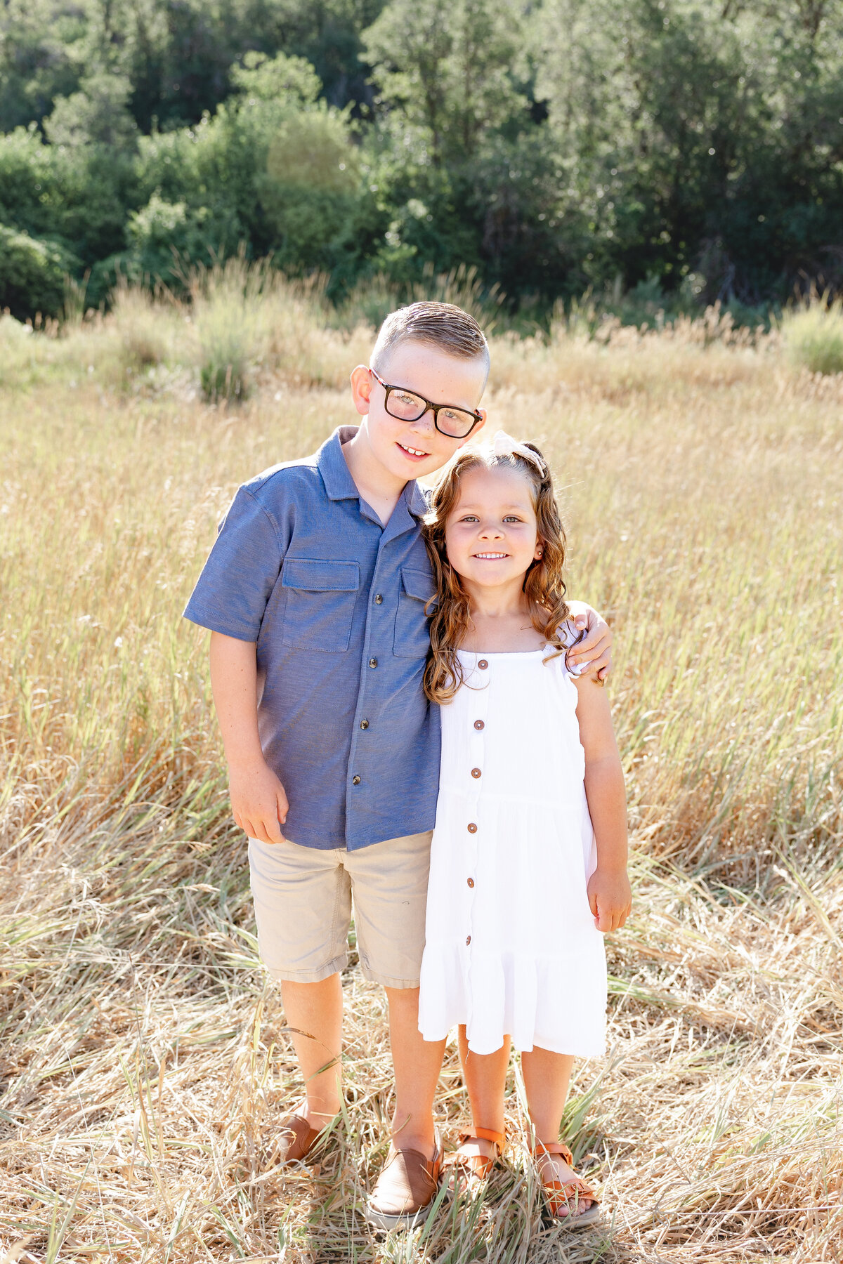 A young brother and sister stand together in a sunlit meadow, smiling as they embrace. Their coordinated outfits and joyful expressions capture a special sibling moment