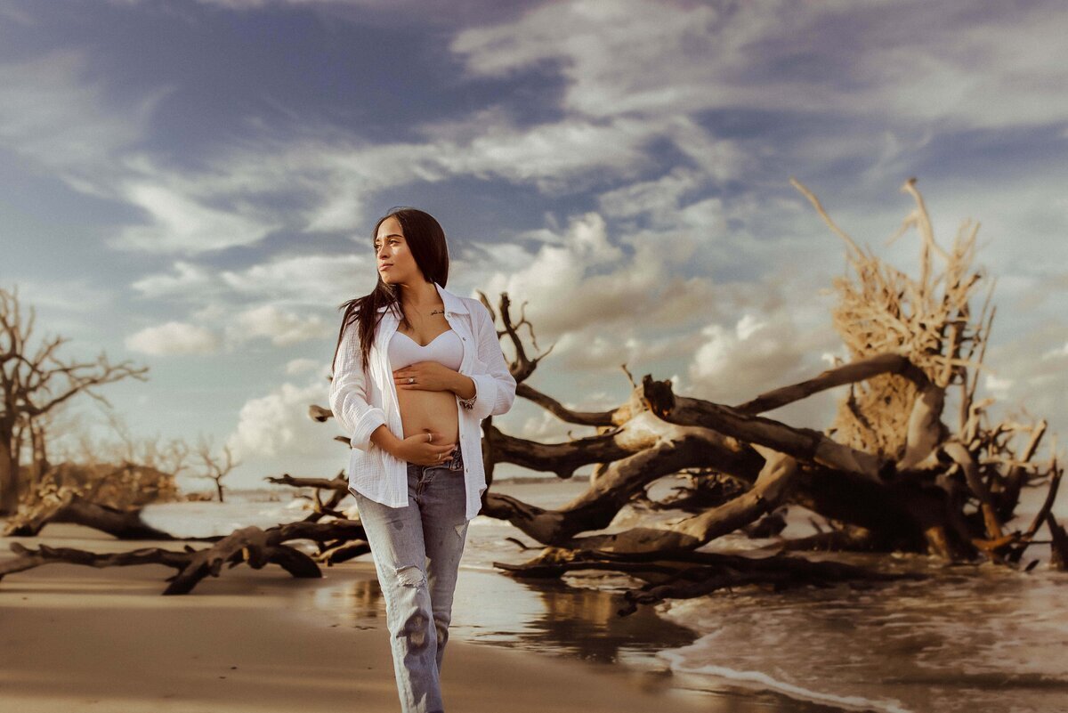 A woman strolls barefoot on wet sand, wearing a white shirt and jeans, cradling her pregnant belly. A backdrop of twisted, leafless driftwood surrounds her serene moment, beautifully captured by a Macon maternity photographer.