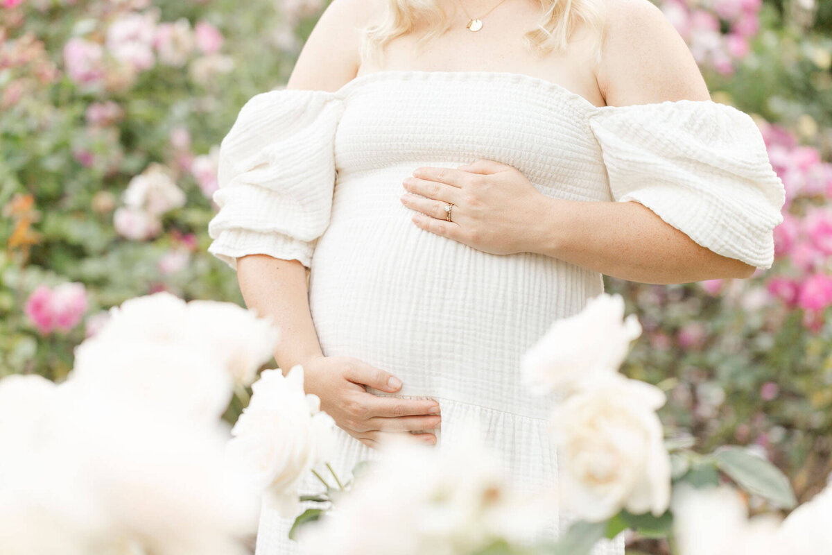 Woman holds pregnant belly in the rose gardens. White roses frame her belly at the bottom of the photo. You cannot see her face in this image.