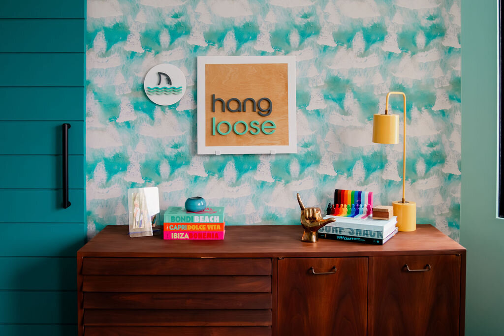 A mid-century modern wooden dresser stands against a wall with a vibrant turquoise and white patterned wallpaper. On the dresser, several stacks of colorful books are arranged. On the left, a stack includes books titled "BONDI BEACH," "CAPRI DOLCE VITA," and "IBIZA BOHEMIA." A small, translucent object and a teal glass ball sit atop this stack. In the center, a shiny gold-colored hand sculpture making the "hang loose" shaka sign rests on a stack featuring a book titled "SURF SHACK." A set of colorful markers is also placed on this stack. A small framed print with the words "hang loose" in teal lettering on a wood-look background hangs on the wall above the dresser, next to a round clock with a shark fin. A slender gold-toned desk lamp with a cylindrical shade stands on the right side of the dresser. To the