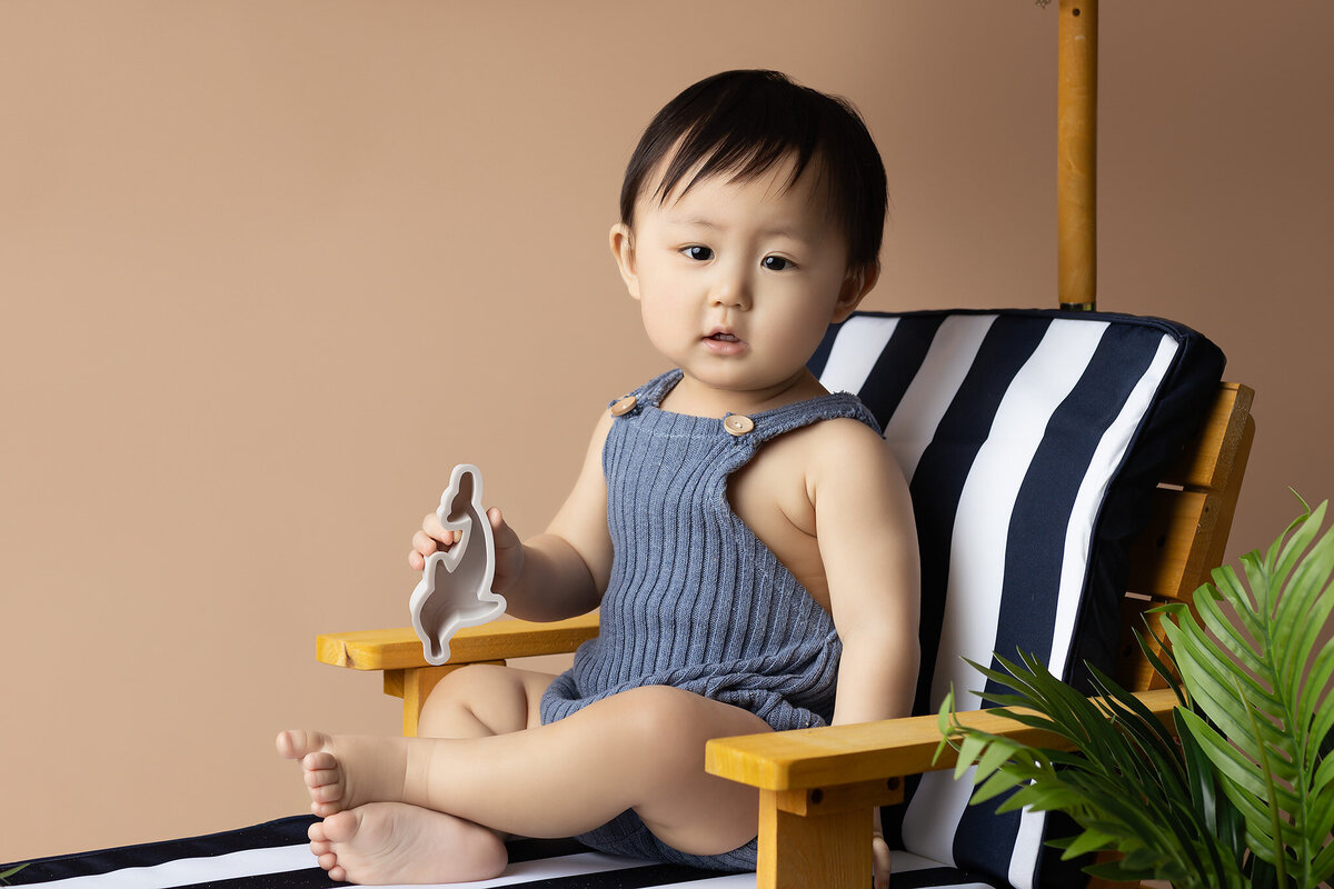 This playful milestone photo captures a baby's beach-themed photoshoot with a striped umbrella and tropical accents. The adorable little one enjoys sitting on a lounge chair while holding a shark toy, bringing a bit of summer fun to the milestone session. Dallas baby milestone photography creates beautiful and unique memories that showcase your child's growth. Experience the creativity of Dallas baby photographers and preserve your little one's first year with expert milestone photography.