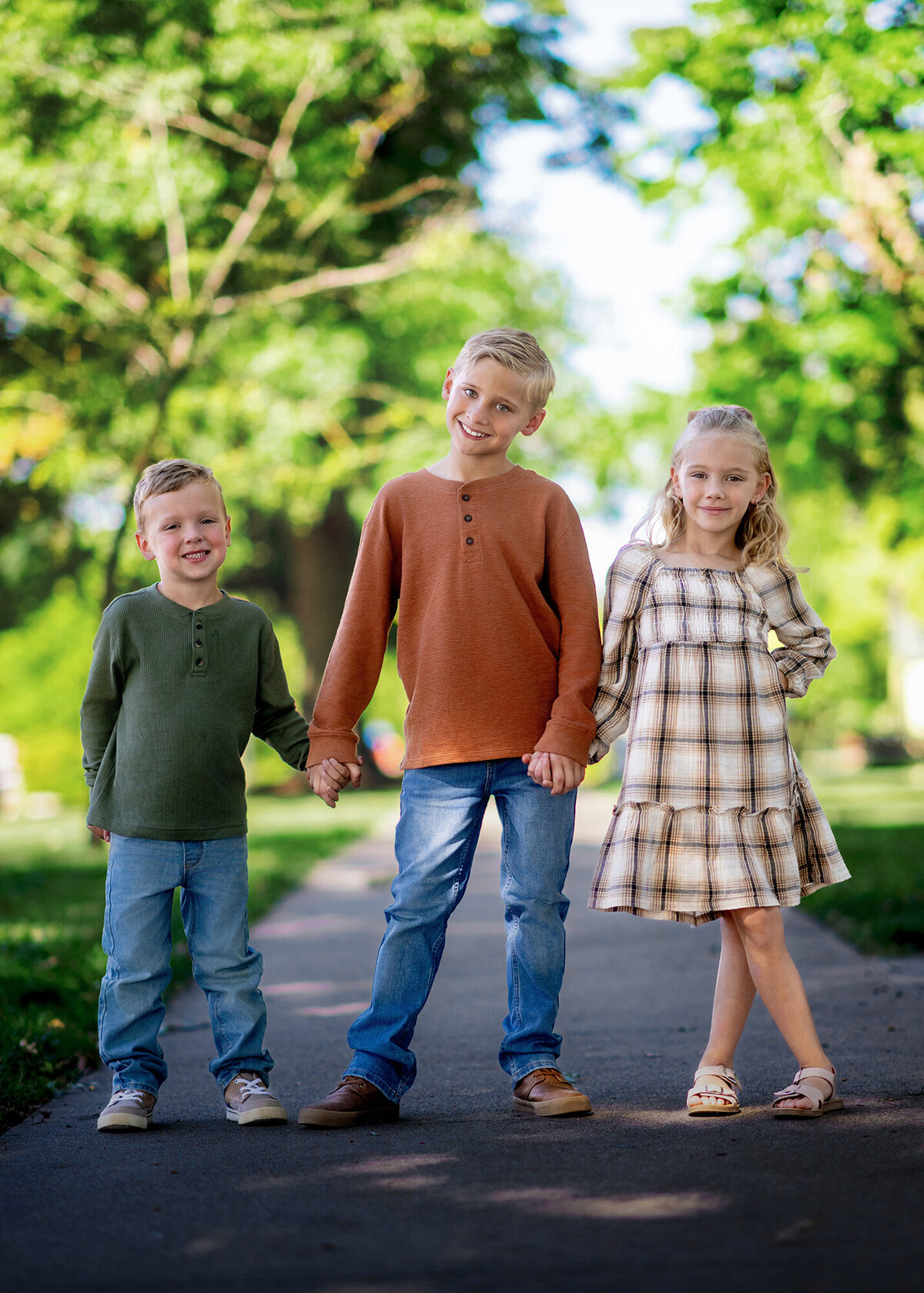 3 siblings standing side by side in a park with trees in the background