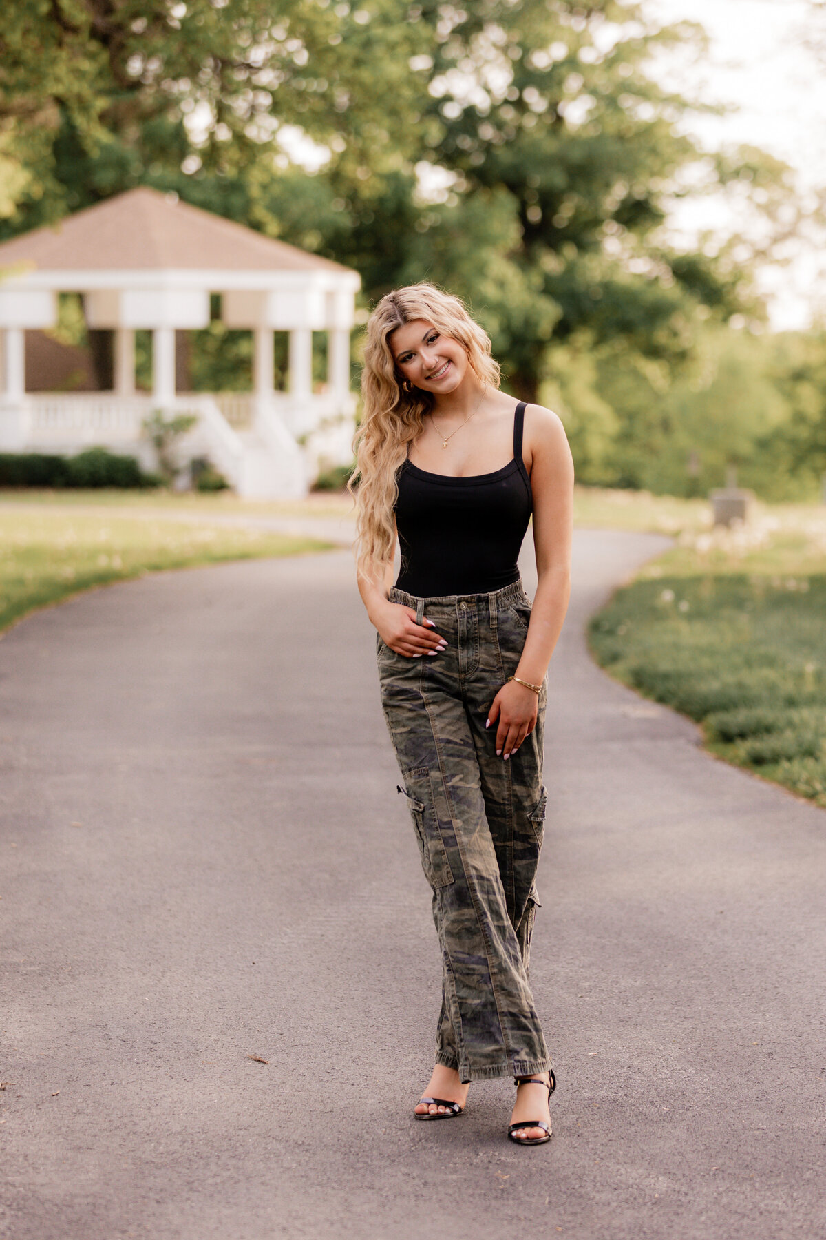 A teen blonde in camoflauge pants poses on a path in front of a gazebo