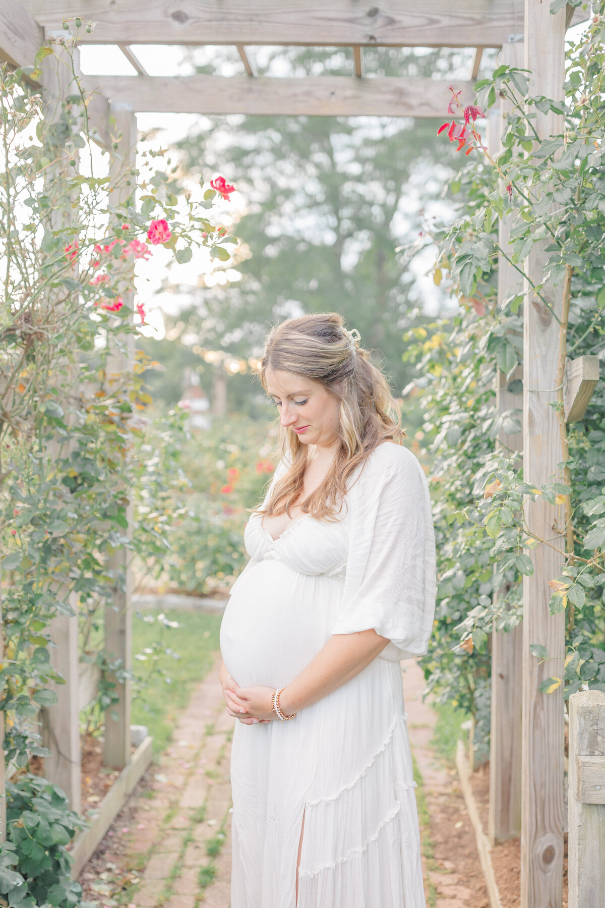 a mother ina  white flowing dress stands holding her pregnant belly and looking down in a serene moment