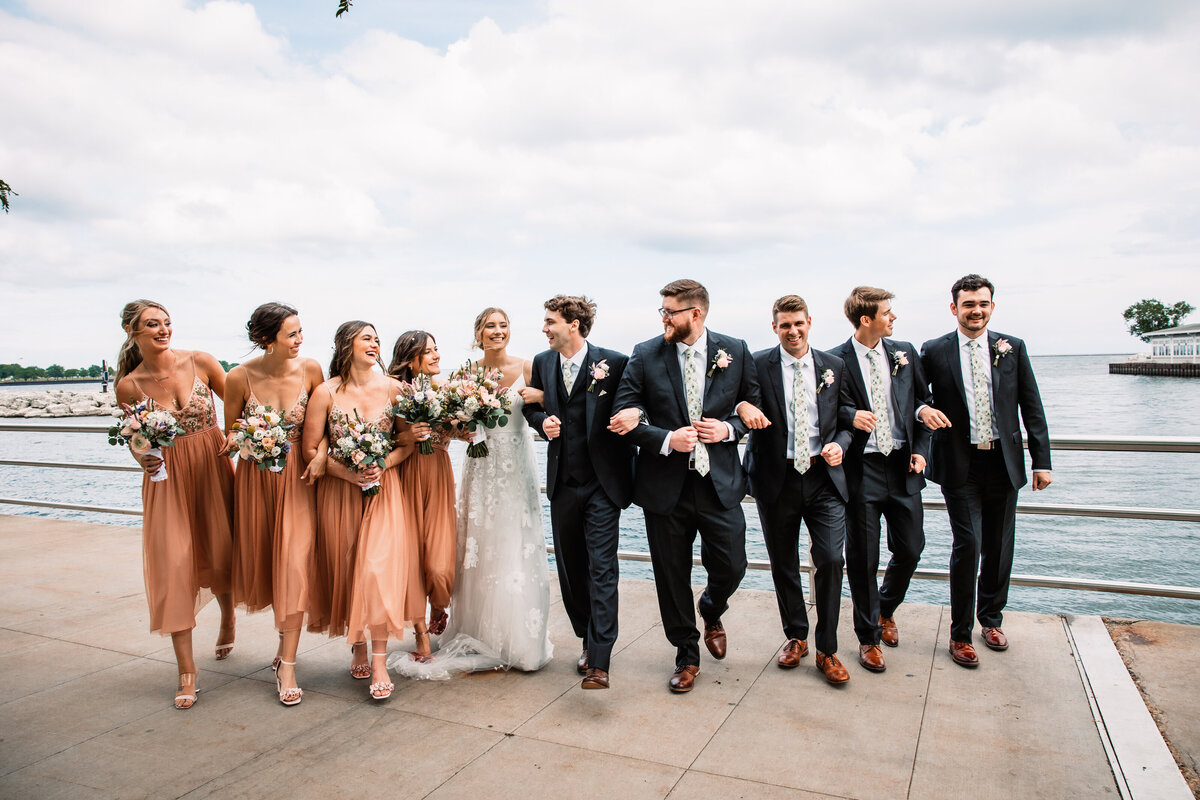 A wedding party and the newlyweds lock arms while walking by the water
