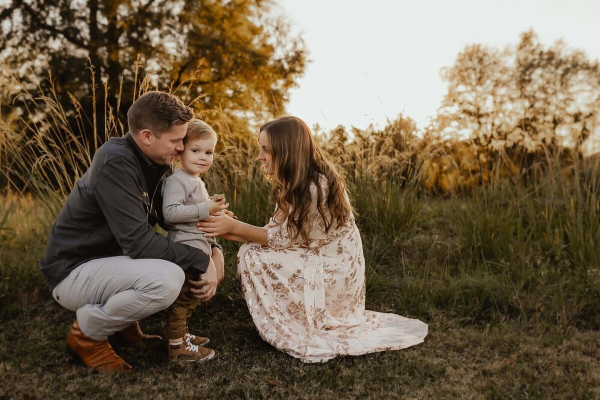 A man, woman, and child crouch together on grass in a field with tall plants and trees in the background, under a warm, golden sky.