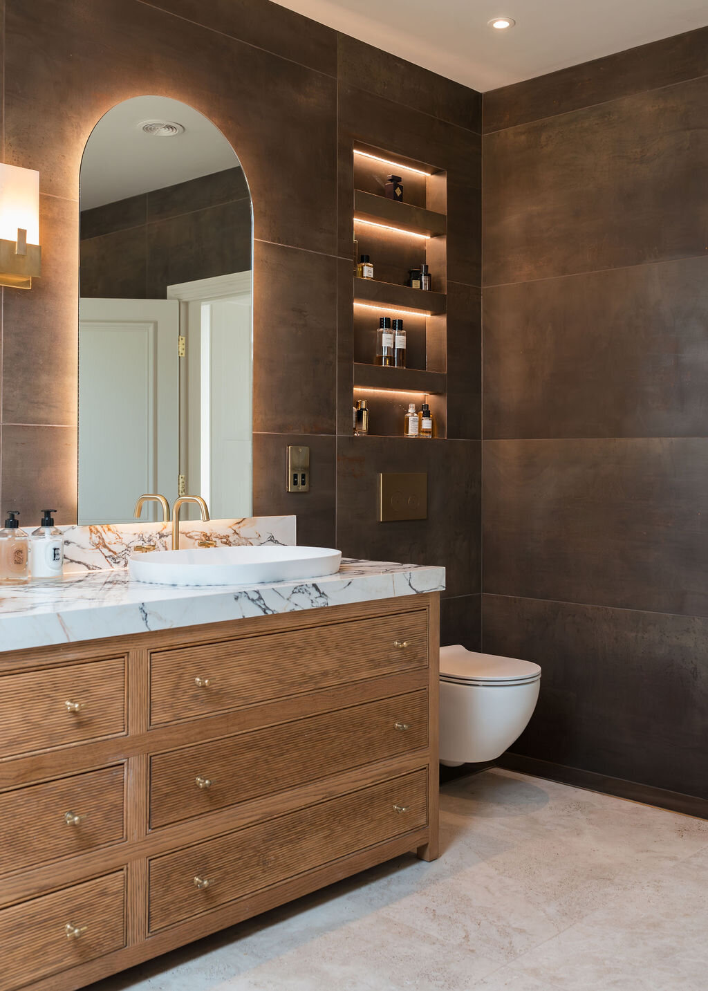 A modern bathroom with a marble countertop and wooden vanity featuring multiple drawers. The wall has dark, large tiles, and there's a gold faucet on the sink. Above the sink, an arched mirror reflects a door. To the left of the sink, sits a white toilet. Above the toilet, lit shelves are built into the wall, emitting a soft light.