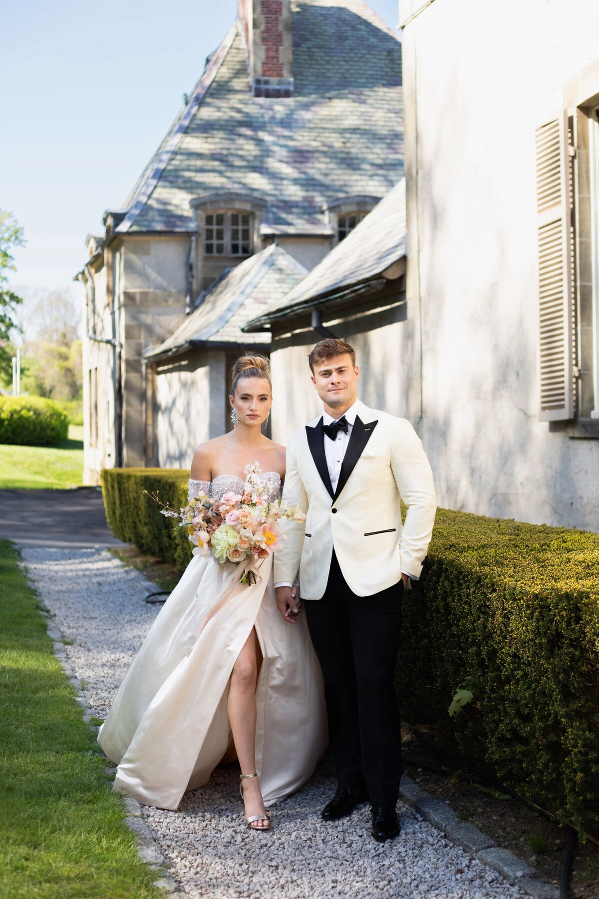 Wedding Photographers in RI | Kelsey Sheehan Photography Timeless Rhode Island Weddings | A couple stands elegantly in evening attire outside a stone building. The woman holds a bouquet of flowers, while the man wears a white tuxedo jacket. Sunlit and serene.