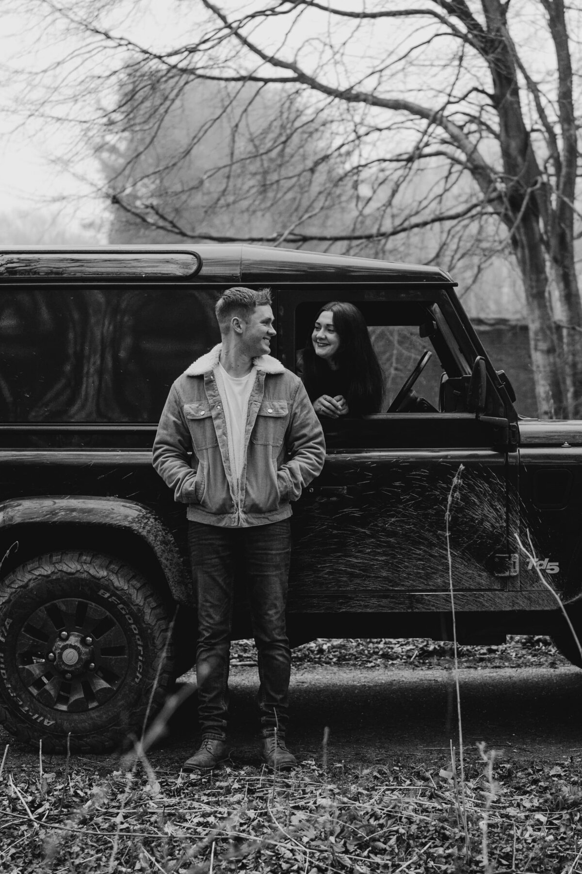 A girl sits in the drivers seat of a Land Rover and hangs her head out of the window to smile at her partner during their engagement photography session.