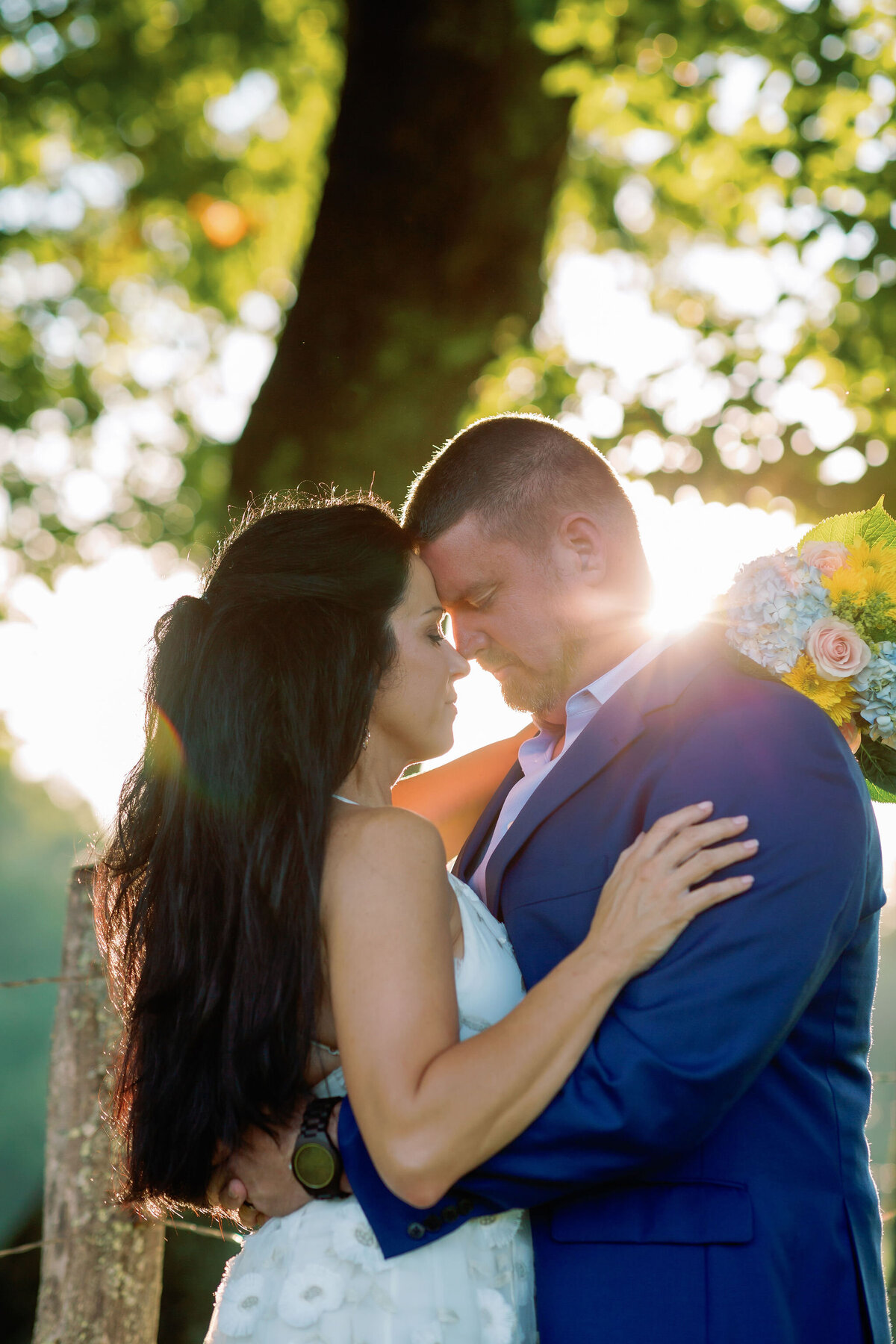 sunsets over the tree line behind the bride and groom who are embracing next to a fence while smiling for their Smoky Mountain elopement in Cades Cove