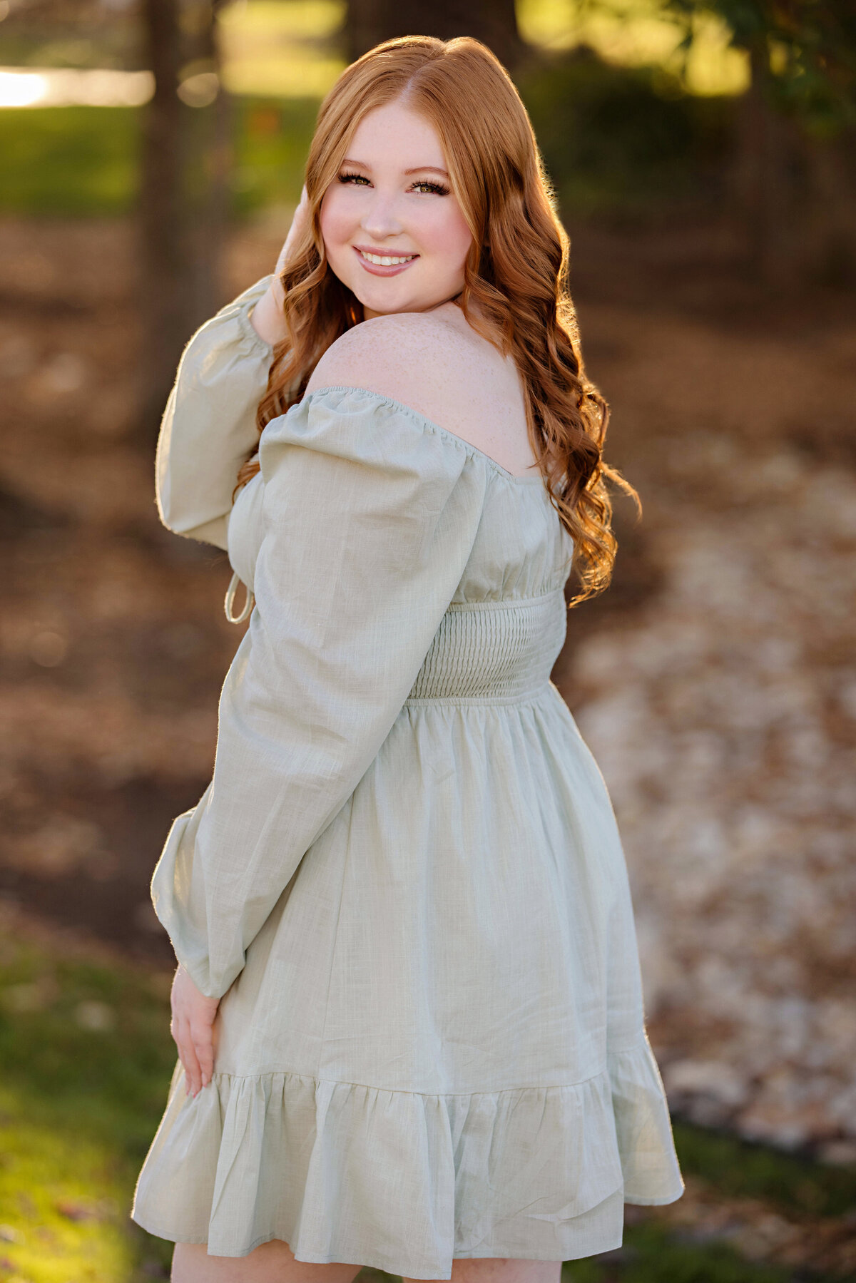 High school senior from South Charlotte wearing a cute sage green dress standing in a wooded area with leaves on the ground and autumn magic hour light through the trees